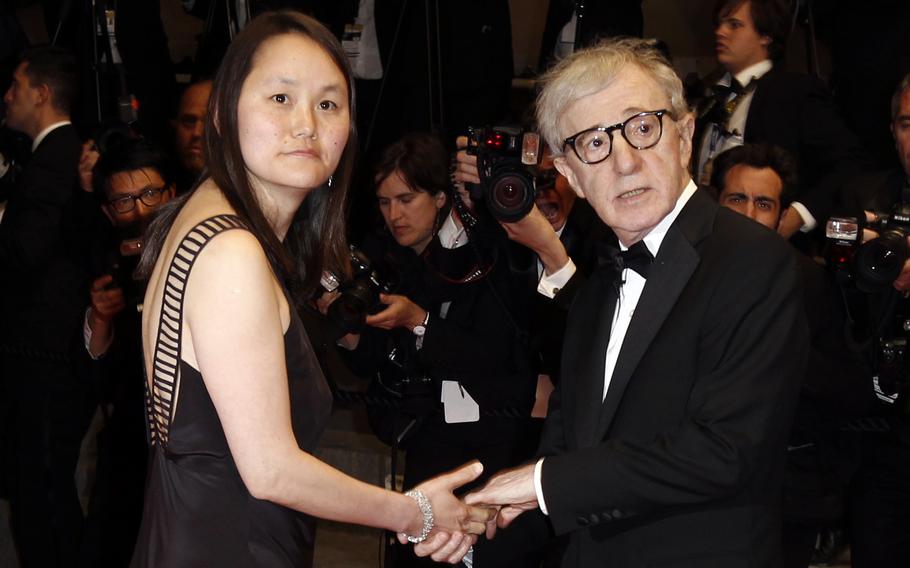 Woody Allen, wearing a tuxedo, holds his wife’s hand as they stand on the red carpet in front of photographers.