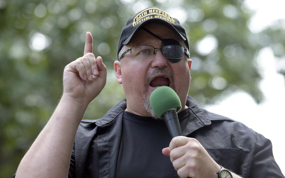 Rhodes raises his right hand, index finger pointing up, and speaks into a microphone at a rally.