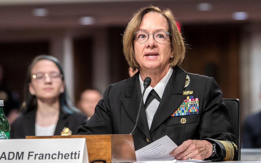 A female Naval officer in black dress uniform sits in front of a gallery of observers during a congressional hearing.