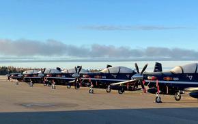 Military jets sit in a row on a runway.