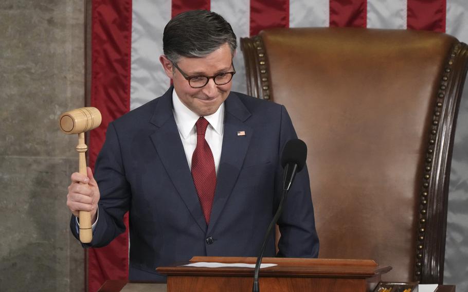 Mike Johnson, wearing a dark suit and glasses, holds up a gavel while standing at a lectern at the head of a Capitol chamber in front of a U.S. flag.