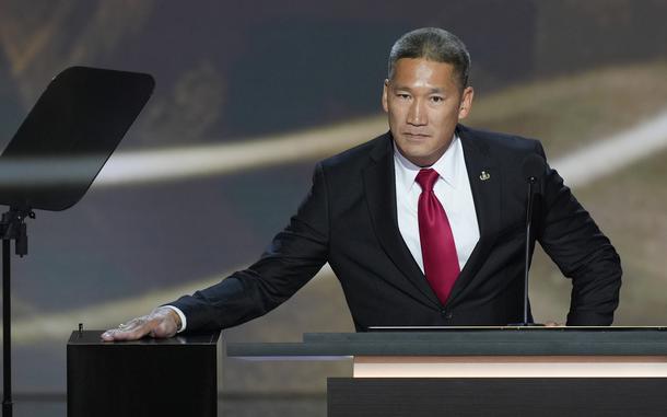 A man in a black suit and red tie leans to one side as he places his hand on a stand.