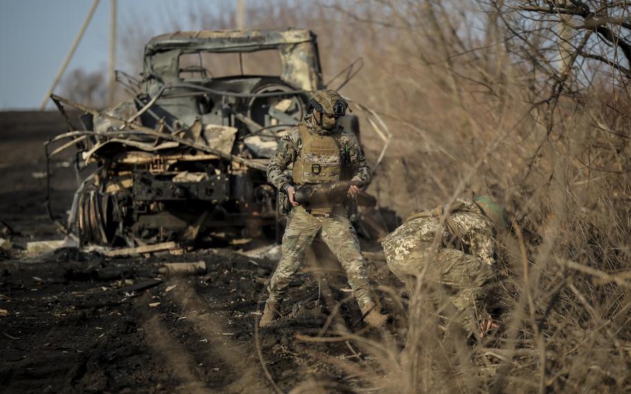 One soldier stands in front of a burned vehicle, while another crouches over the floor.