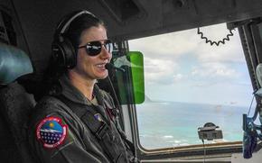 A woman in a flight suit and sunglasses wearing a radio headset sits in an airplane with a view of an ocean outside the window.