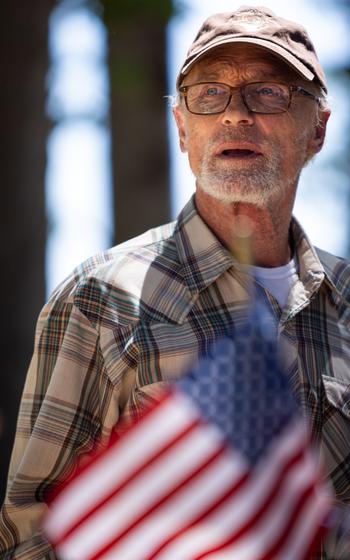 A man in a checkered flannel and hat, with an out-of-focus American flag in front of him.