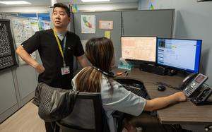 A California Poison Control System male employee stands next to a woman sitting in front of a computer with a headset around her neck. She is working on the poison control hotline at the system’s San Francisco office, which is located in a hospital.