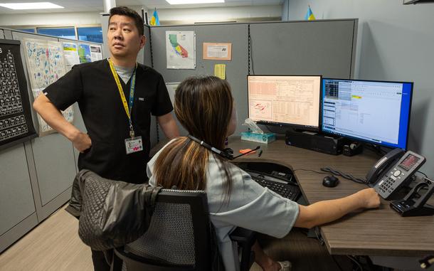 A California Poison Control System male employee stands next to a woman sitting in front of a computer with a headset around her neck. She is working on the poison control hotline at the system’s San Francisco office, which is located in a hospital.