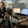 A California Poison Control System male employee stands next to a woman sitting in front of a computer with a headset around her neck. She is working on the poison control hotline at the system’s San Francisco office, which is located in a hospital.