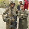 A woman in combat gear stands next to an Afghan boy.