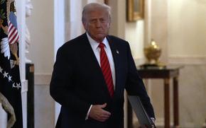 Donald Trump, wearing a dark suit and red tie walks down a hallway in the White House while carrying a binder in his left hand.