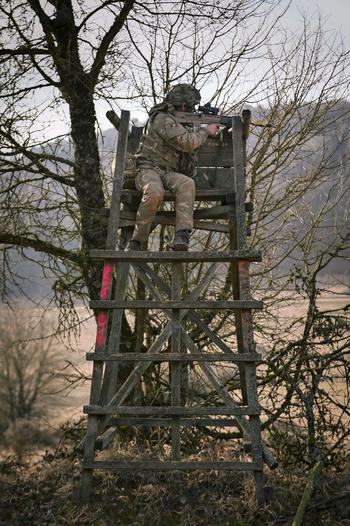 A U.K. soldier points a rifle from on top of a wooden lookout.