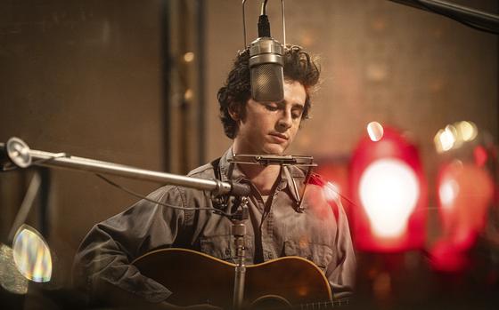 Timothée Chalamet, playing Bob Dylan, in front of a microphone and holding a guitar. 