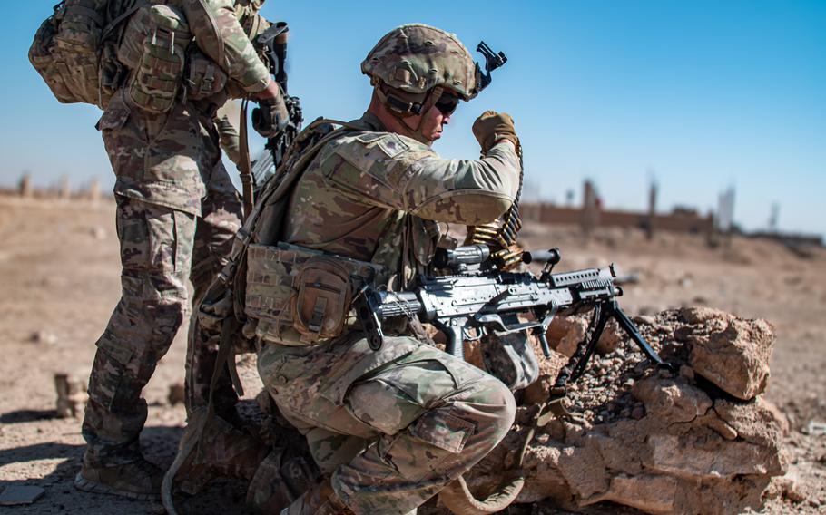 A soldier in combat gear kneels while holding a machine gun in a desert setting.