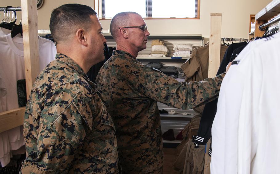 Two Marines in camouflage uniforms look through a rack of clothing on hangers inside a store.