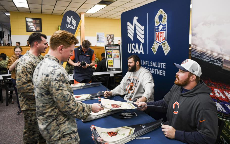 Two football players in street clothing sitting at a booth while two service members stand in front of them.