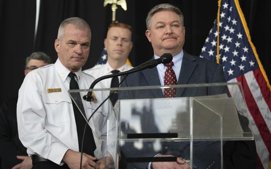 A man in a dark suit stands at a podium next to a man in a shirt and tie at a press conference.