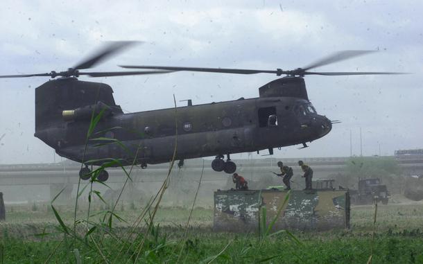 An Army helicopter hovers over three small soldiers standing on a large block.