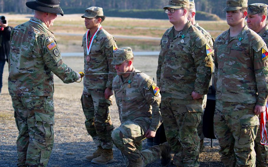 A U.S. sergeant, surrounded by other soldiers, kneels and is knighted with a sword.