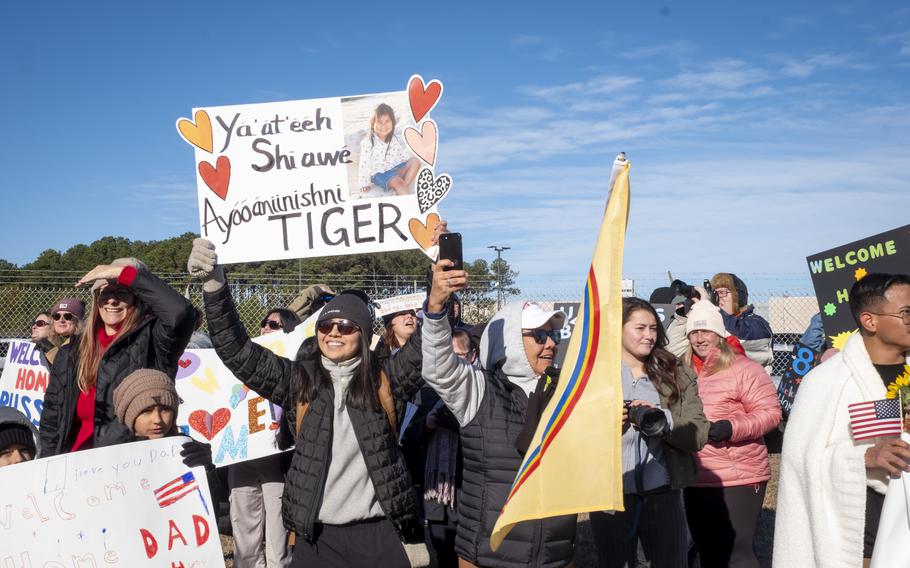 A woman, waiting for sailors among a group of friends and family, holds up a sign that says sign that says, “Ya’at’eeh Shi awe. Ayooanunishini Tiger.”