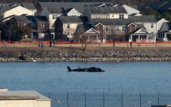 Wreckage from an airplane and helicopter crash floats in a river with houses along the shore line in the background.