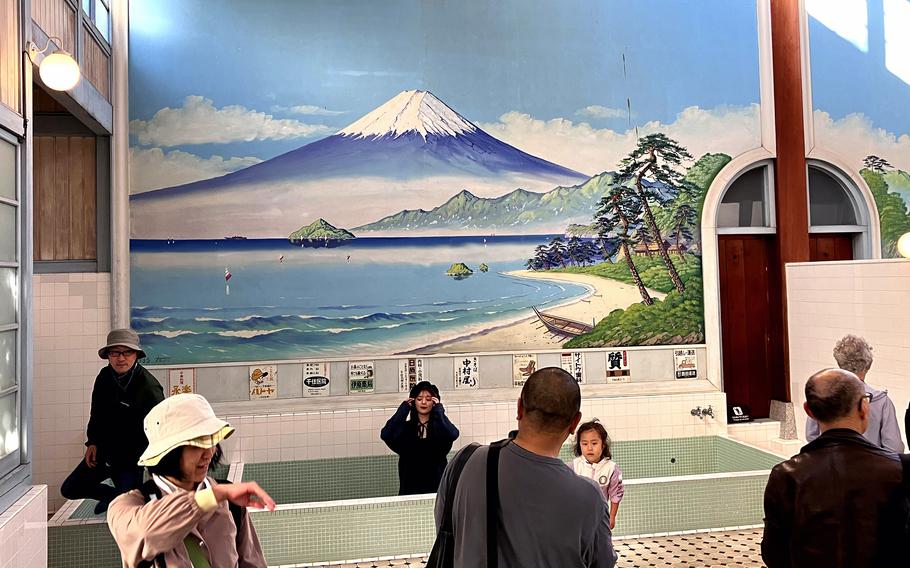 A mural showing a painting of Mount Fuji and a lake adorns a wall with people milling around in front of it.