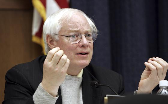 A man wearing a black suit jacket and a gray sweater sits and gestures with both hands in front of him.