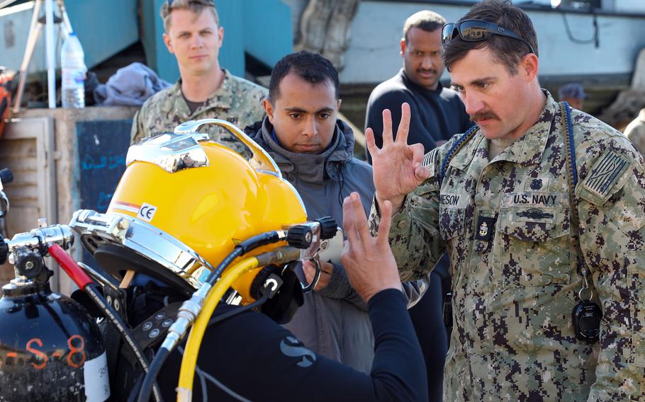 A U.S. Navy sailor gives the ‘OK’ sign to a man in a diving suit and yellow helmet.