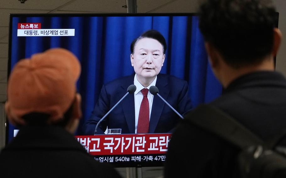 Two men, seen from behind, watch a television screen broadcasting remarks by South Korean President Yoon Suk Yeol.