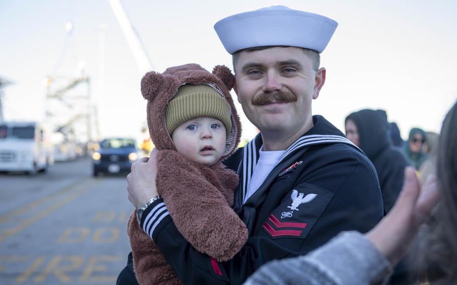 A sailor embraces his child, who is wearing a bear outfit.