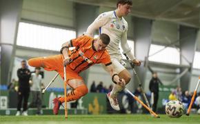 Two soccer players on different teams, each with crutches, in mid-air as a soccer ball is seen in front of them.