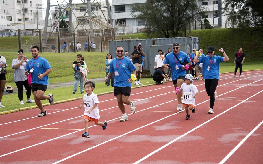 Children in white shirts race on a track while adults in blue run behind them and cheer.