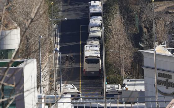 A road lined with buses is seen just beyond the top of a security barrier.