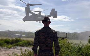 A man stands in front of an MV-22B Osprey as it lands.