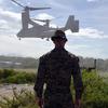 A man stands in front of an MV-22B Osprey as it lands.