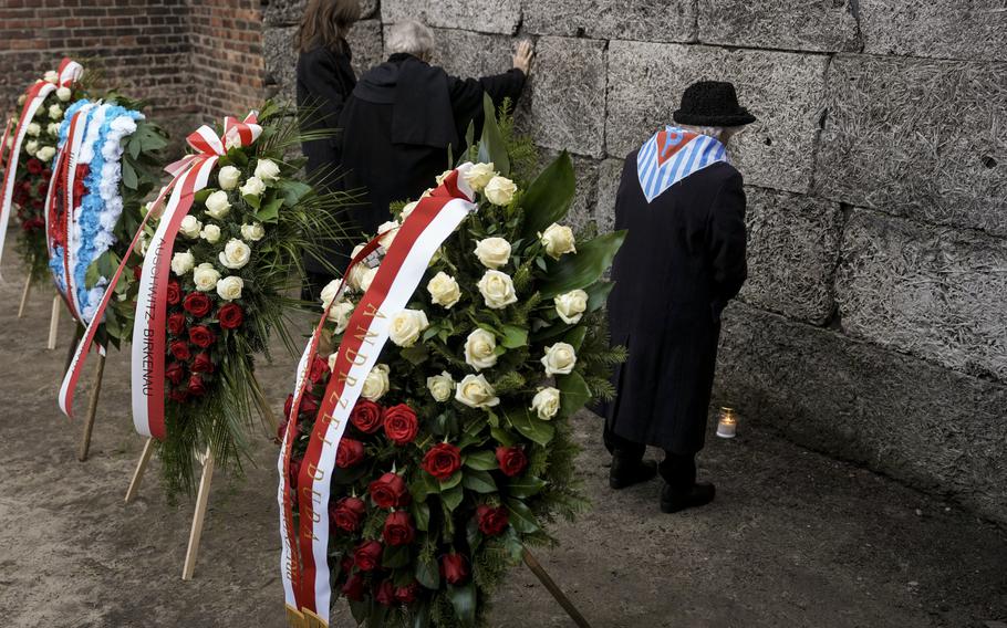 Elderly Auschwitz survivors, some wearing blue-and-white striped scarves, stand together at the Death Wall.