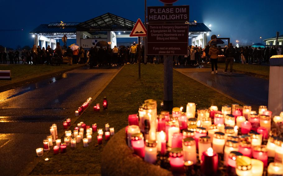 Candles placed on a stone planter and the ground  glow in the foreground as people protest at the closed main gate of Spangdahlem Air Base on a rainy evening. 