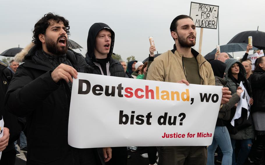 Protesters hold a sign that translates to “Germany, where are you?” Another sign in the background says, “prison for murder.” Some protesters hold umbrellas and candles.