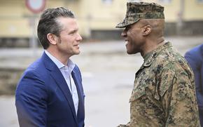 Pete Hegseth, wearing a dark blazer, shakes hands with a military general in camouflage uniform and hat.