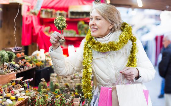 A cheerful woman adorned in a cozy white jacket and shimmering gold garland examines a charming miniature tree. With bags in hand, she embodies the spirit of festive shopping, surrounded by an array of handcrafted ornaments and joyful decorations that fill the air with delight.