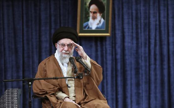 Iran’s supreme leader raises one hand in salute while sitting against the backdrop of a blue curtain.