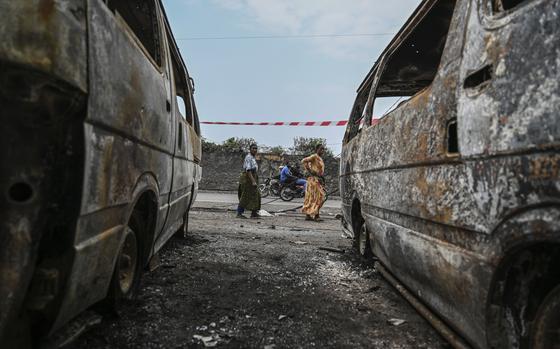 Two people are seen walking through the gap between two destroyed cars in the foreground.