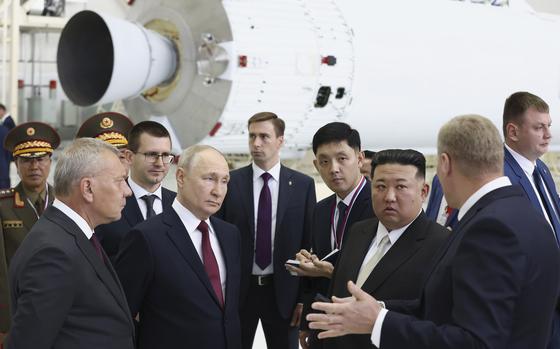 Five men in suits stand in a semi-circle and talk in front of a piece of a rocket and some military officials in uniform.