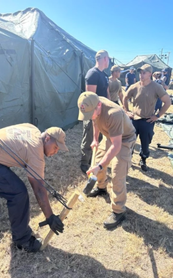 Two sailors setting up a tent by driving a wooden stake into the ground.