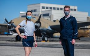 An Air Force airman wearing a navy blue shorts and a gray T-shirt stands next to a fellow airman in a navy blue track suit. 