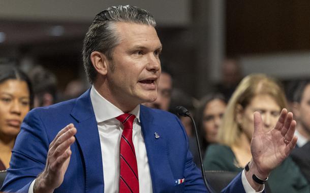 Pete Hegseth raises both his hands while speaking into a microphone at his seat.