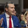 Pete Hegseth raises both his hands while speaking into a microphone at his seat.