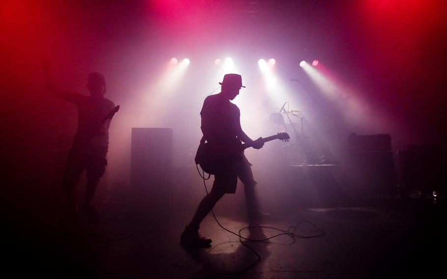 A man in a hat and suit jacket playing guitar is silhouetted against spotlights in the background.