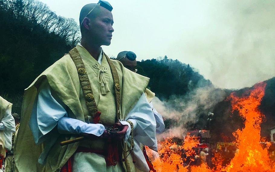 A monk stands in the foreground while a fire rages behind him.