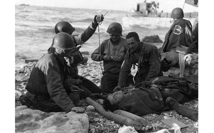 Nurses administer a plasma transfusion to a wounded survivor of a landing craft at “Fox Green” sector portion of Omaha Beach. The photograph shows how promptly medical treatment was administered on the beach. Note the inflatable life belt used as a pillow, and the feet of second prostrate man on (right). The flag on the far right is a flag of the Red Cross. The soldiers are from the 16th Infantry Regiment, 1st Infantry Division. The exact date of the image is unknown. Both June 6 (D-Day) and as June 12 are used.