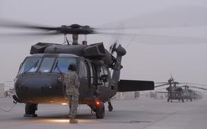 A soldier in camouflage uniform and black helmet stands next to a helicopter on a tarmac.